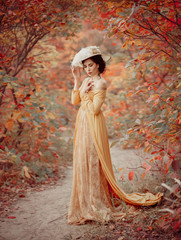A young brunette woman with an elegant, hairstyle in a hat with a strass feathers. Lady in a yellow vintage dress walks through the autumn landscape. Artistic portrait