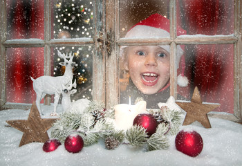Kind schaut freudig überrascht aus dem weihnachtlich dekorierten Weihnachtsfenster, innen leuchtet...