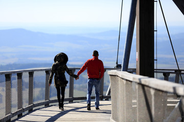 Kobieta i mężczyzna trzyma się za ręce idąc po platformie widokowej w górach. - obrazy, fototapety, plakaty
