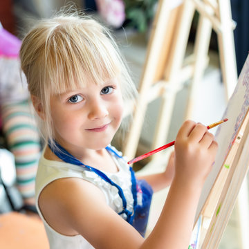 Cute blond smiling girl painting on easel in workshop lesson at art studio. Kid holding brush in hand and having fun drawing with paints. Child development concept