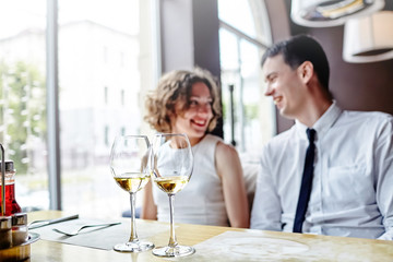 Two glasses of white wine standing on a wooden table in a restaurant. In the background a laughing couple in love. Selective focus
