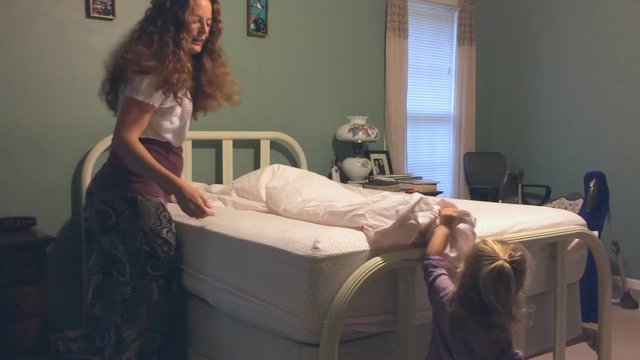 Mother making bed with little daughter together in bedroom