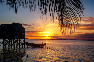 An overwater shack is silhouetted by a dramatic orange sunset and framed by palm fronds on the tiny Caribbean Island of Tobacco Caye in Belize's barrier reef