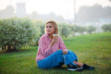Handsome blonde woman in stylish blouse and jeans sitting on the green grass in the park and drinking coffee.