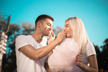 Happy young cheerful loving couple walking outdoors in the amusement park posing eat sweeties cotton candy floss.