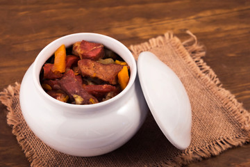 Meat stew with vegetables, stewed in oven in ceramic white pot on linen napkin, wooden background.