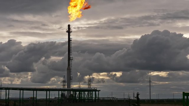 A tall GAS TORCH emits a bright flame into a cloudy evening sky at an oil facility. Industrial scene with heavy atmosphere and dramatic contrast.