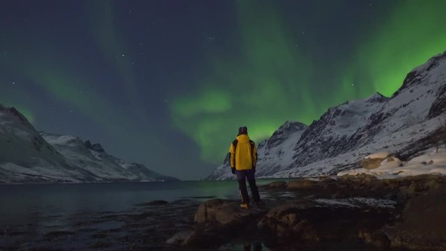 Northern Lights (Aurora Borealis) filmed in real time. Two tourists photographers take pictures and enjoy the calm display of green and purple colours, surrounded by mountains and arctic fjord