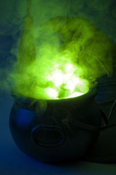 Happy halloween, alchemy and magic spell potion concept with a pot or calderon boiling a green liquid with fumes and smoke coming out, next to a broomstick and a witch or wizard hat