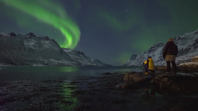 Northern Lights (Aurora Borealis) filmed in real time. Two tourists photographers take pictures and enjoy the calm display of green and purple colours, surrounded by mountains and arctic fjord