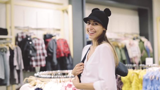 a young joyful smiling girl trying on a funny hat and backpack in a store and posing in front of the mirror