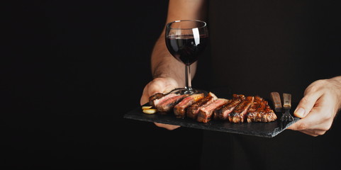 Man holding juicy grilled beef steak with spices and red wine glass on a stone cutting board on a...