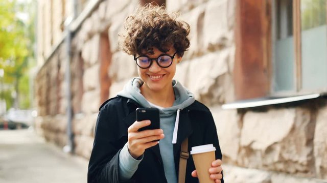 Smiling young woman in glasses is using smartphone looking at screen while walking outdoors in city with to-go coffee. Youth lifestyle, street and technology concept.