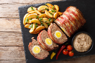 Delicious sliced meat loaf wrapped in bacon with potato wedges and sauce close-up on a slate board....