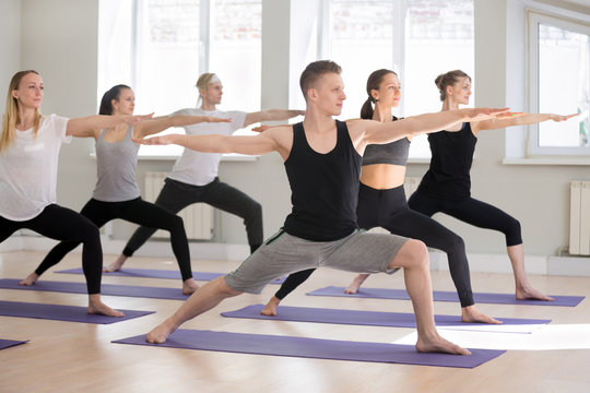 Group of young attractive sporty people practicing yoga lesson, doing Warrior Two exercise, Virabhadrasana II pose, working out indoor full length, students training in club studio. Well-being concept