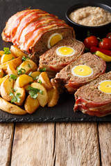 Delicious sliced meat loaf wrapped in bacon with potato wedges and sauce close-up on a slate board. vertical