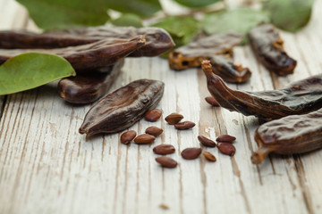 Carob pods with seeds and green leaves on white wooden table. Healthy eating, food background.