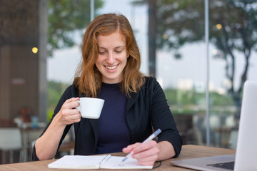 Positive student reading copybook during coffee break. Young woman in dark blue jacket drinking coffee and writing notes in notebook. Studying anywhere concept - obrazy, fototapety, plakaty
