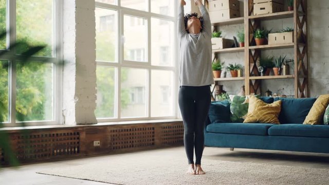 Pretty girl in sports clothing is doing yoga at home then resting with hands in Namaste. Stylish loft style interior with large window and modern furniture is visible.