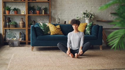 Slender young woman is bending forward to one leg then relaxing in Cobbler's pose during yoga practice at home. Wellness, youth and sports concept.