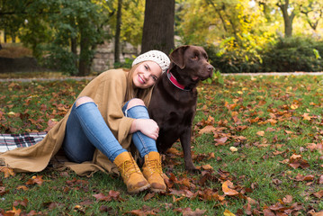 Fototapeta premium A beautiful young blond-haired woman is having fun with her big brown labrador dog in the park, in a pile of autumnal fallen leaves