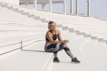 Middle Eastern Girl with short braided hair sitting on the stairs laughing on a construction site wearing gray and black fitness outfit on a hot bright sunny day.   