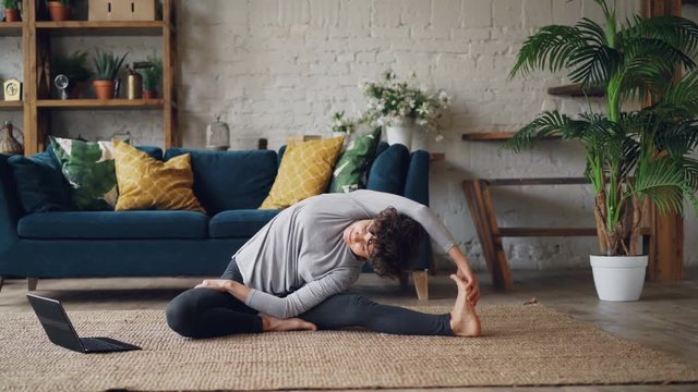 Beautiful girl is watching yoga tutorials on laptop screen and exercising sitting on floor at home alone. Modern technology, self-development and youth concept.