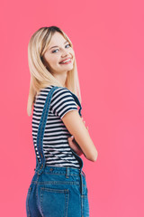 smiling beautiful girl in denim overall standing with crossed arms and looking at camera isolated on pink