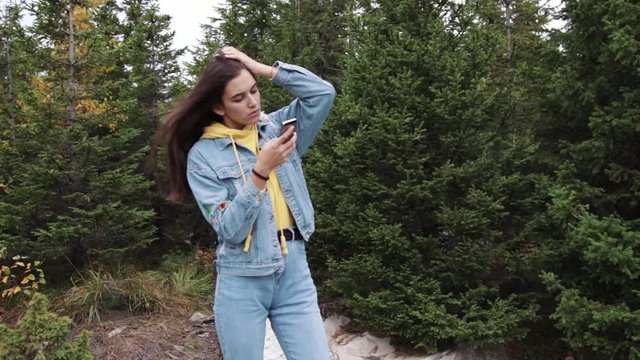 Young teen coed using touch screen smartphone while in nature. Hipster using social media and smiling while reading mobile. Beautiful woman in denim jacket in spruce forest