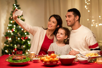 holidays, family and celebration concept - happy mother, father and little daughter having christmas dinner and taking selfie picture by smartphone at home