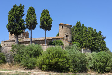 Fototapeta premium Spain, Catalonia, Peratallada: Panorama side view of famous Sant Esteve church of the small fortified medieval Spanish town with green trees, beautiful old s rutted stone streets and blue sky.