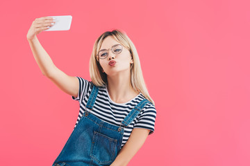 portrait of young woman with duck face taking selfie on smartphone isolated on pink