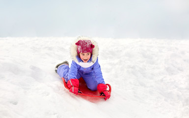 childhood, sledging and season concept - happy little girl sliding down hill on snow saucer sled outdoors in winter