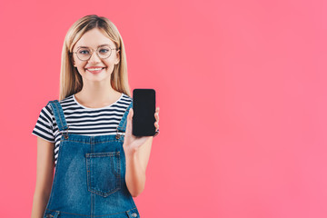 Fototapeta premium portrait of smiling woman in eyeglasses showing smartphone with blank screen isolated on pink