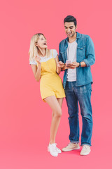 cheerful young man showing smartphone to laughing girlfriend isolated on pink