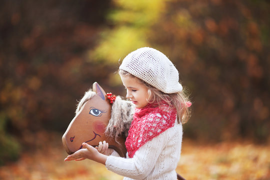 Cute little girl feeds a wooden toy horse in the autumn garden.