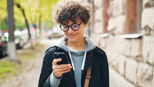Slow motion of smiling woman in glasses and trendy clothing using smartphone looking at screen and walking outdoors in city. Youth lifestyle, street and technology concept.