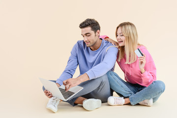smiling young couple with credit card using laptop isolated on beige