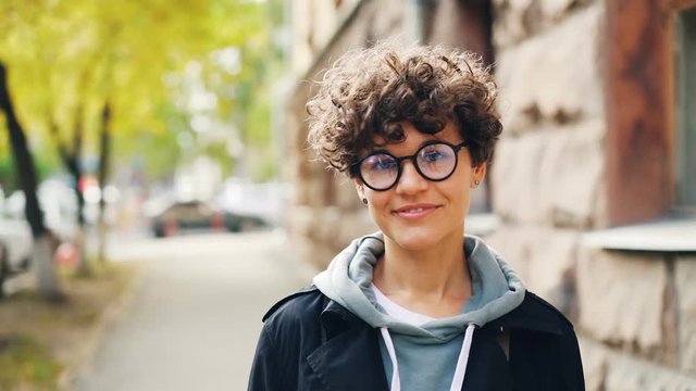 Slow motion portrait of attractive lady wearing glasses and casual clothing smiling and looking at camera standing outdoors in the street. Youth, emotions and city concept.