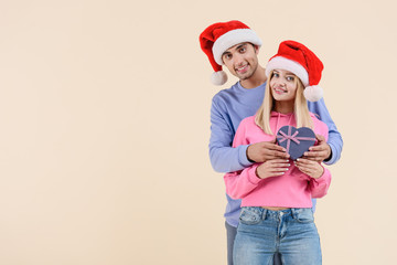 happy couple in santa hats holding heart shaped present and smiling at camera isolated on beige