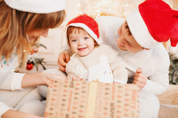 Merry Christmas and Happy Holidays! Cheerful parents and their cute daughter girls exchanging gifts. Parent and one little children having fun and playing together near Christmas tree indoors.