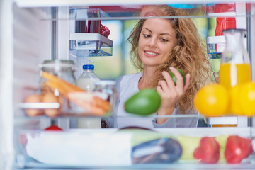 Woman standing in front of opened fridge and taking avocado. Fridge full of groceries. Picture taken from the iside of fridge.