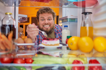 Man eating cheesecake. Unhealthy eating concept. Picture taken from the inside of fridge full of...