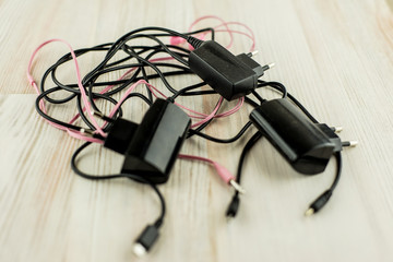 the need for universal battery charger: black 3 different chargers with different connectors and earphones tangled cables together, on the white oak table, in short focus