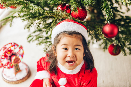 Christmas portrait of adorable 3 year old asian toddler girl wearing red Santa dress and hat, sitting on the floor, top view