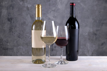 Two bottles of red and white wine with matte black & white blank labels on concrete wall background. Wineglasses half full with vintage expensive wine of different type. Close up, copy space.