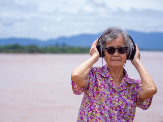Portrait of happy senior woman listening music on headphone for relaxing with side the river. Relaxing time concept.