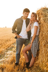 Full length photo of young couple man and woman walking on field, and standing near big haystack