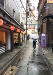 man with umbrella walking on street puddles in rainy day on red lights background. Travel to Japan, Kyoto. Street Bokeh Lights Out Of Focus.