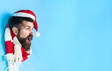 Surprised man looking through hole in blue paper. Bearded man in Santa hat& scarf making hole in...